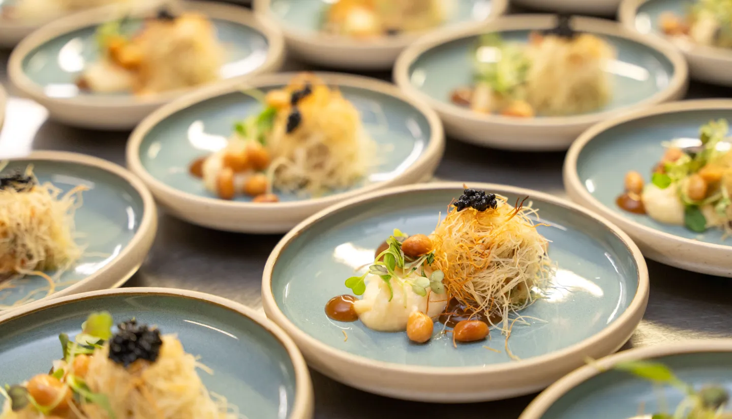 Elegantly plated dishes featuring a main course of crispy shredded pastry, topped with caviar, microgreens, and small colorful garnishes, served on light blue ceramic plates for a refined presentation.